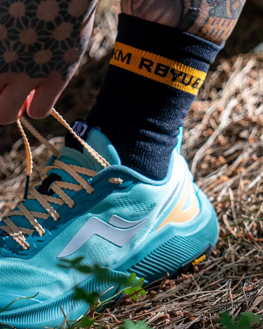 The black RBYU sock with its amber RBYU cuff band rising out of a teal trail shoe on a bed of pine needles.