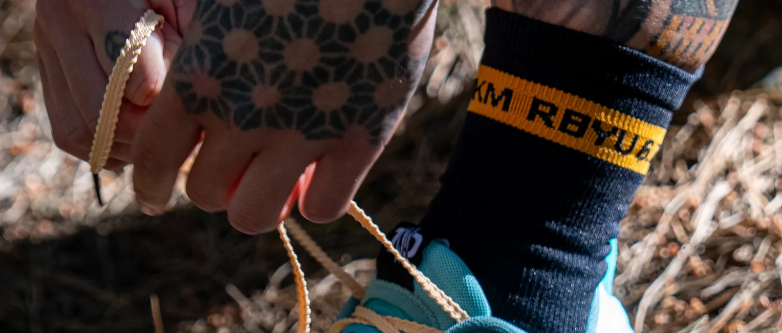 Tattooed hands pulling trail shoe laces tight beside the amber RBYU 6.7KM cuff band of the sock.