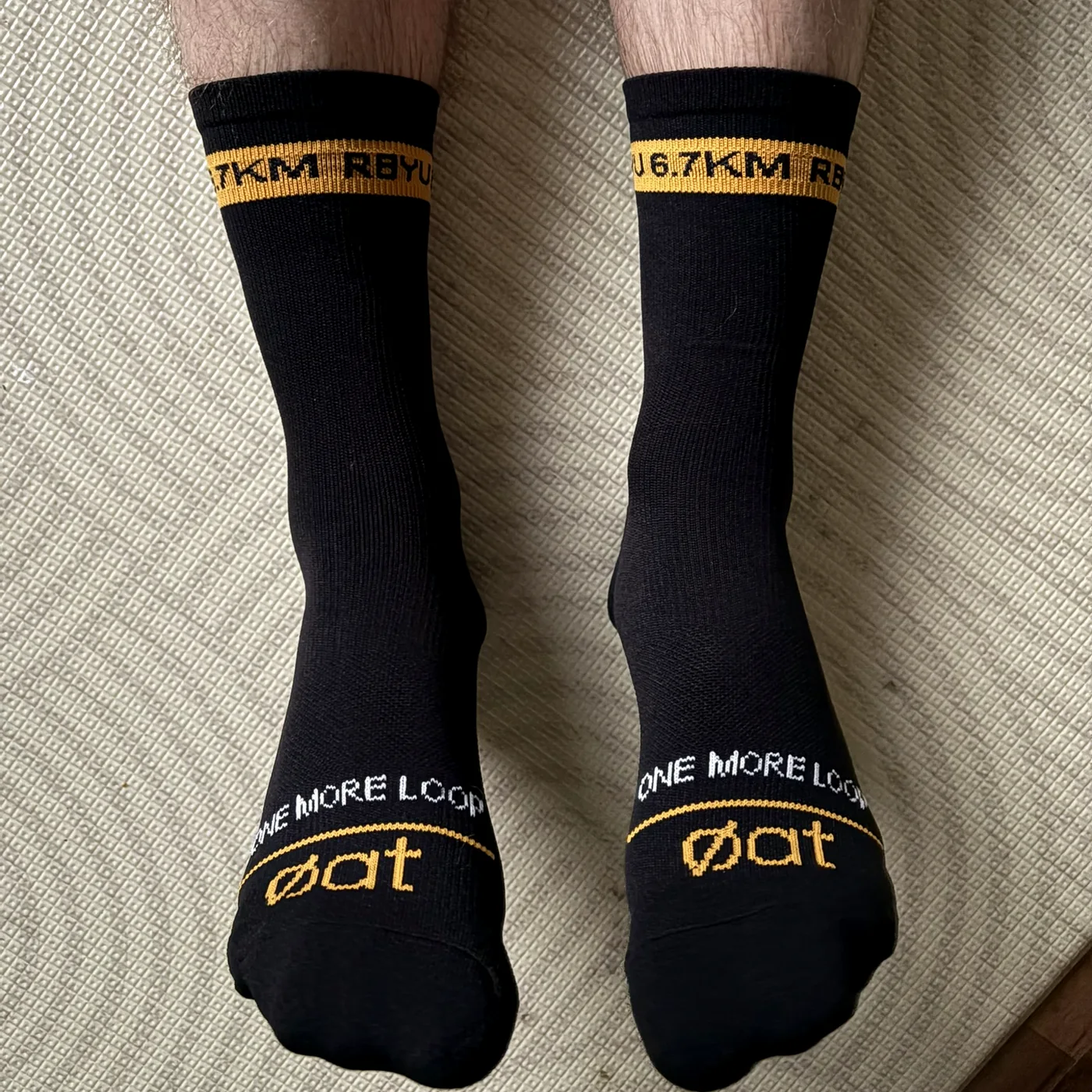Both RBYU socks worn and seen from above, showing the amber RBYU 6.7KM cuff band, the white ONE MORE LOOP lettering and the gold øat toe mark.