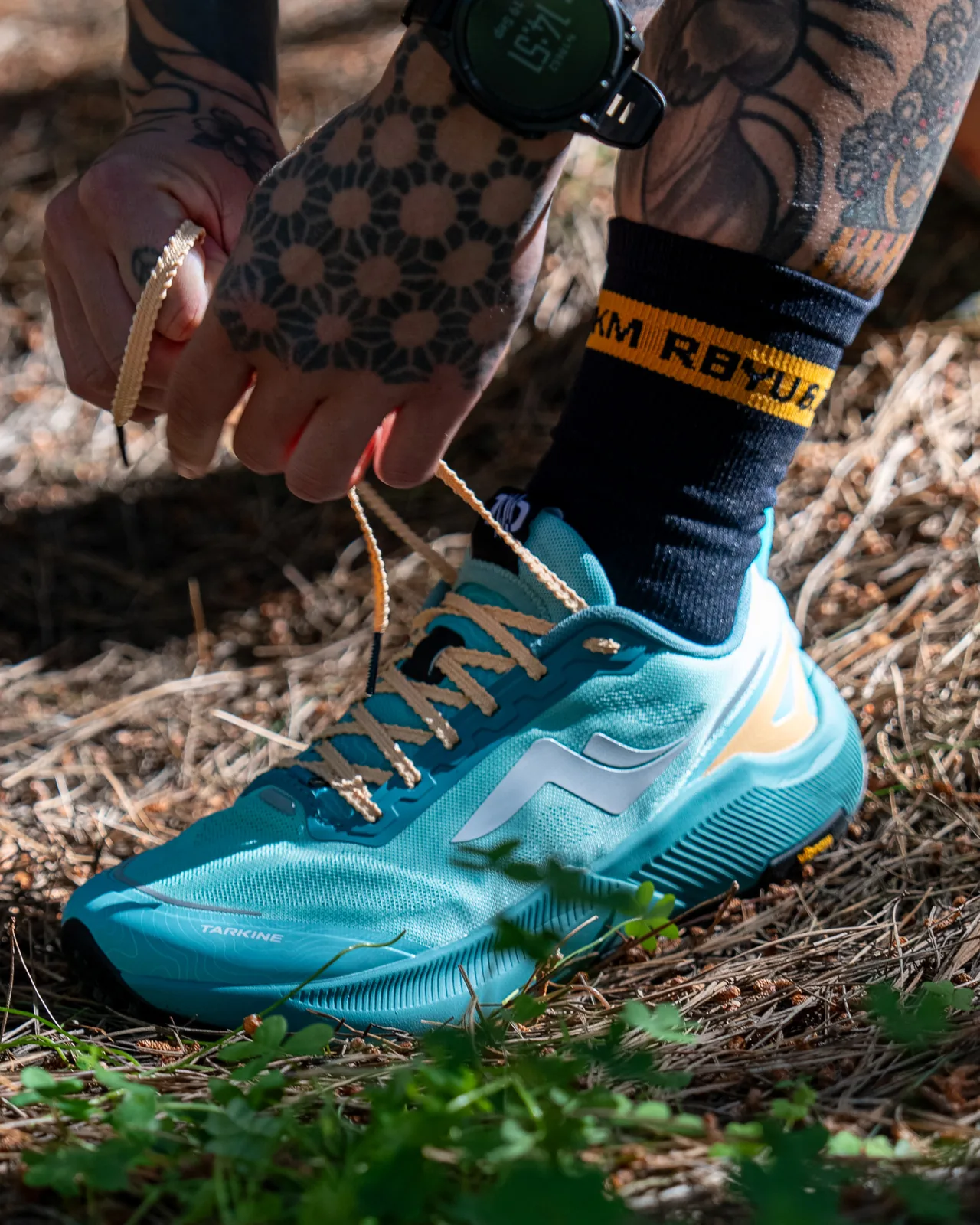 A runner lacing a trail shoe on a pine needle track, the black RBYU sock showing its amber RBYU 6.7KM cuff band above the shoe.