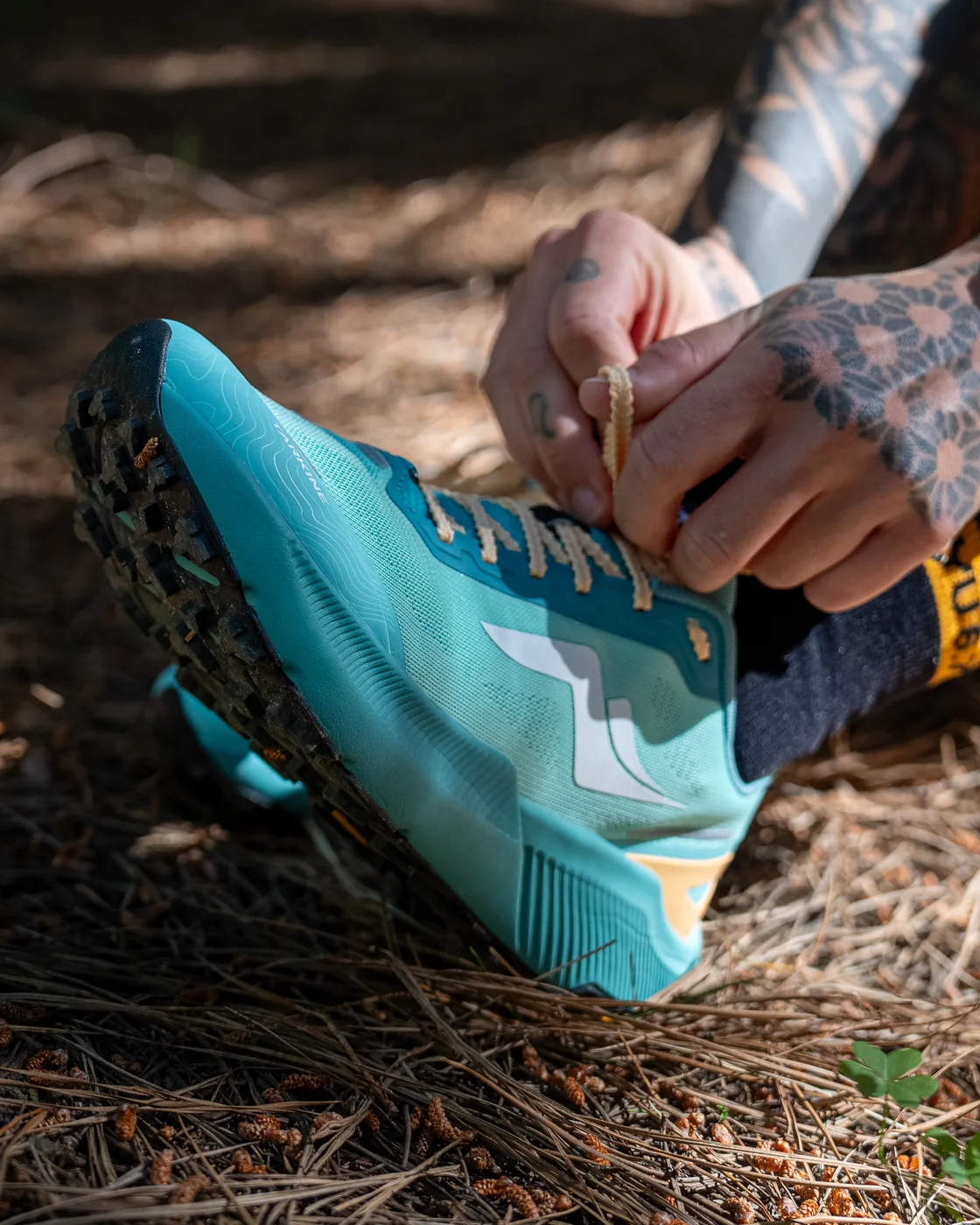 A runner lacing a teal trail shoe on a pine needle track, the black sock and amber cuff at the ankle.